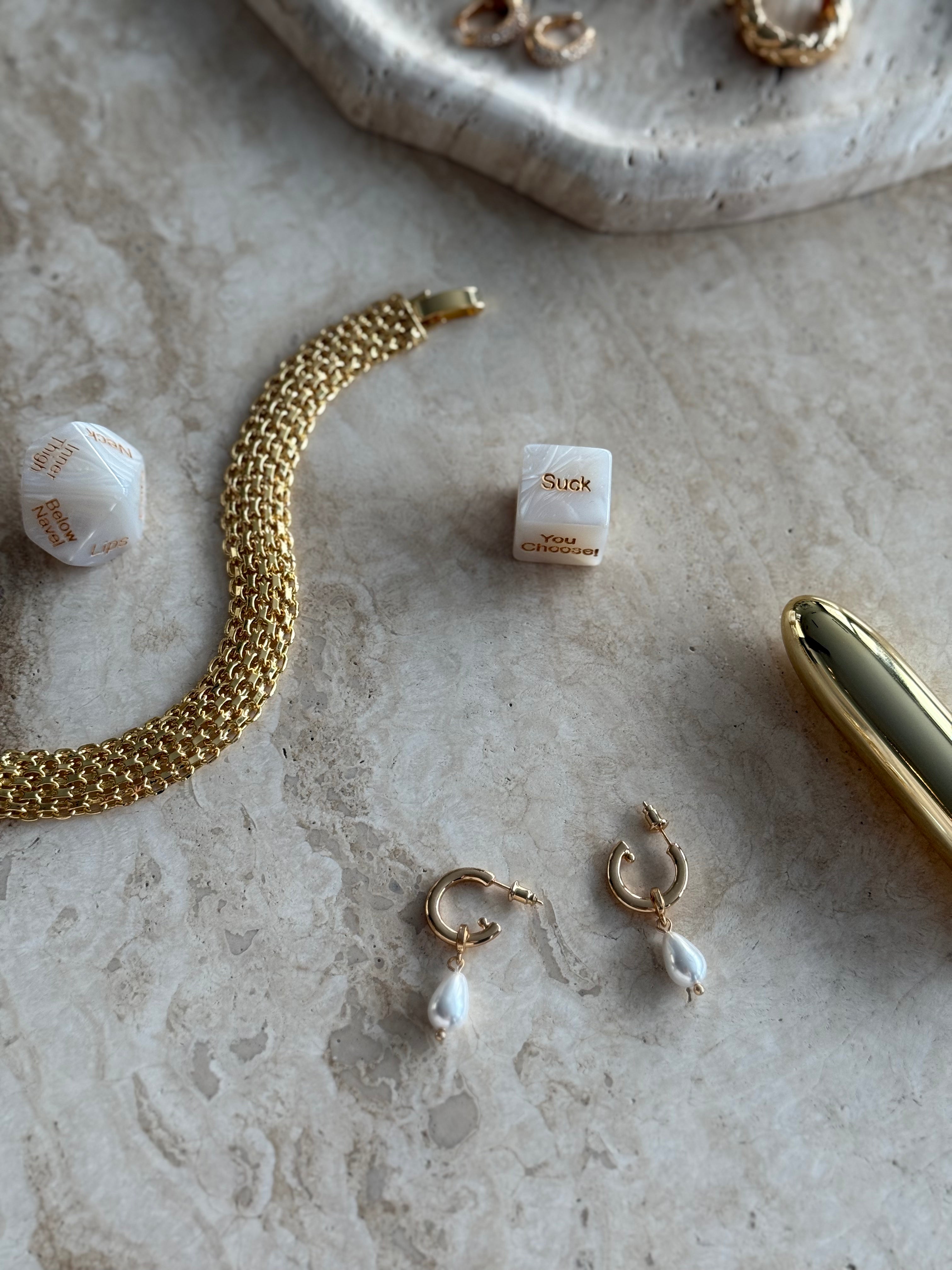 The Candace Drop Pearl Earrings in gold styled on beige marble surface beside gold jewelry and sensual dice by SELF Seductions