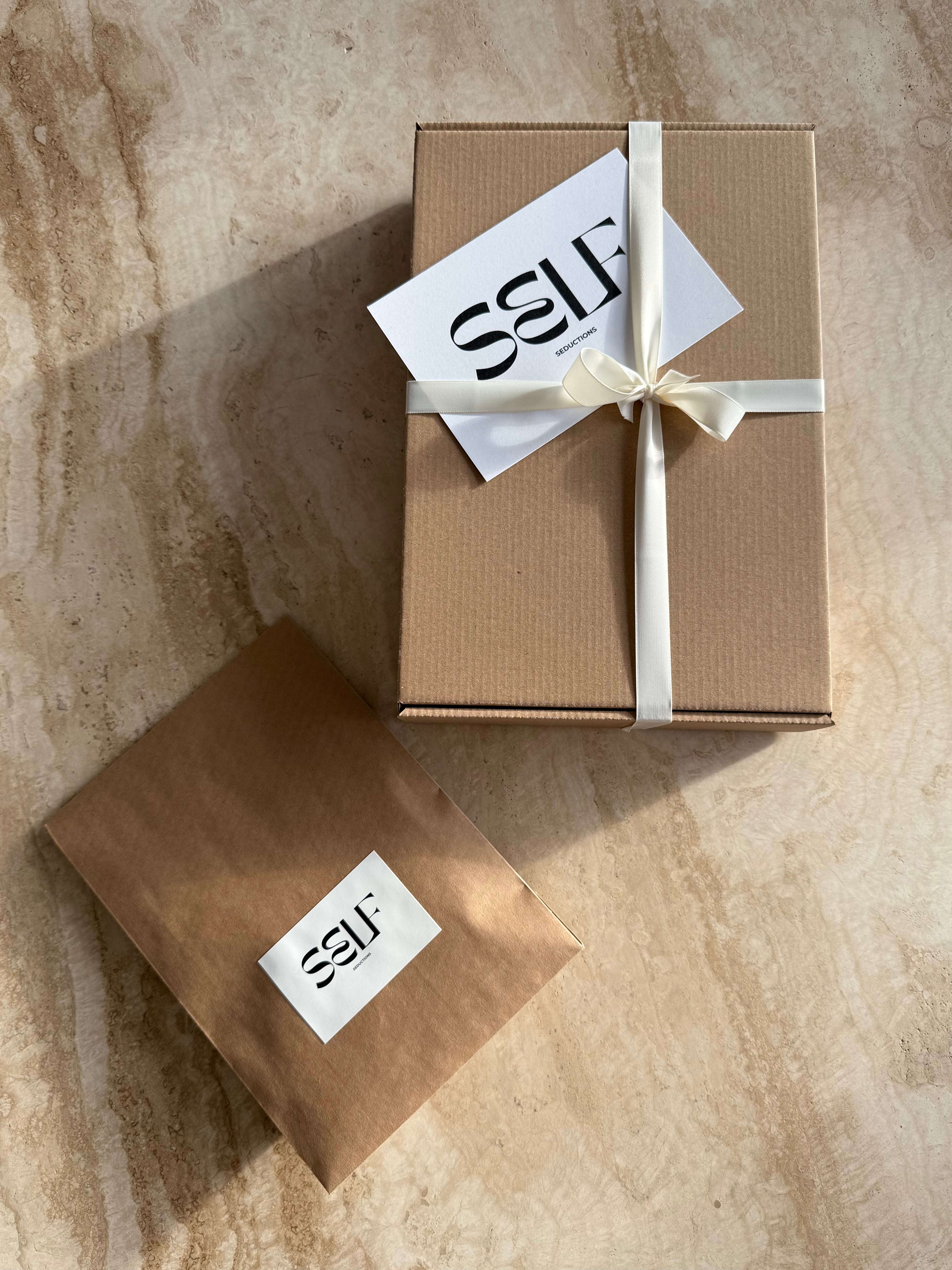 Luxury eco packaging by SELF Seductions – brown mailer with white brand label and tied ribbon on stone surface