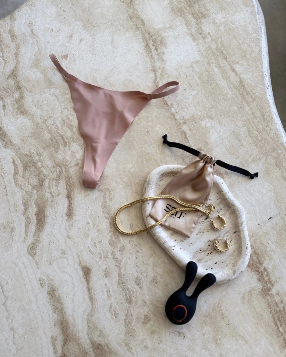 The Kylie Skin Yoga Thong in nude beige – soft seamless thong with gold jewelry on marble surface – bold yet minimal lingerie still life by SELF Seductions