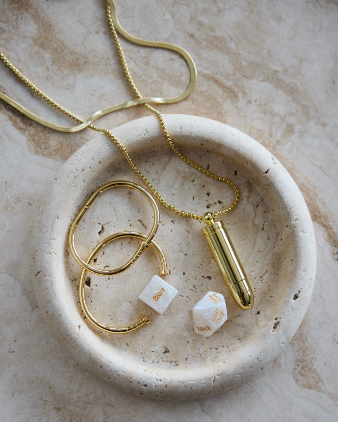 Lolita Undercover gold bullet necklace styled on beige marble surface with gold jewelry and sensual dice by SELF Seductions.