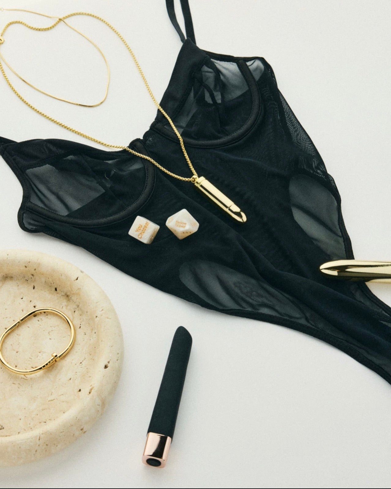 The Lolita Undercover Necklace discreet wearable massager in gold, styled with a black mesh bodysuit, silicone massager and intimacy dice on a clean white background.
