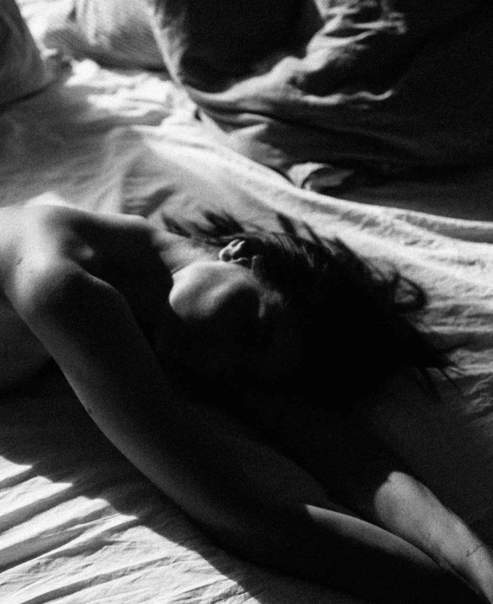 Black and white photo of a woman lying on a bed with soft shadows, part of the SELF Seductions intimate wellness aesthetic.