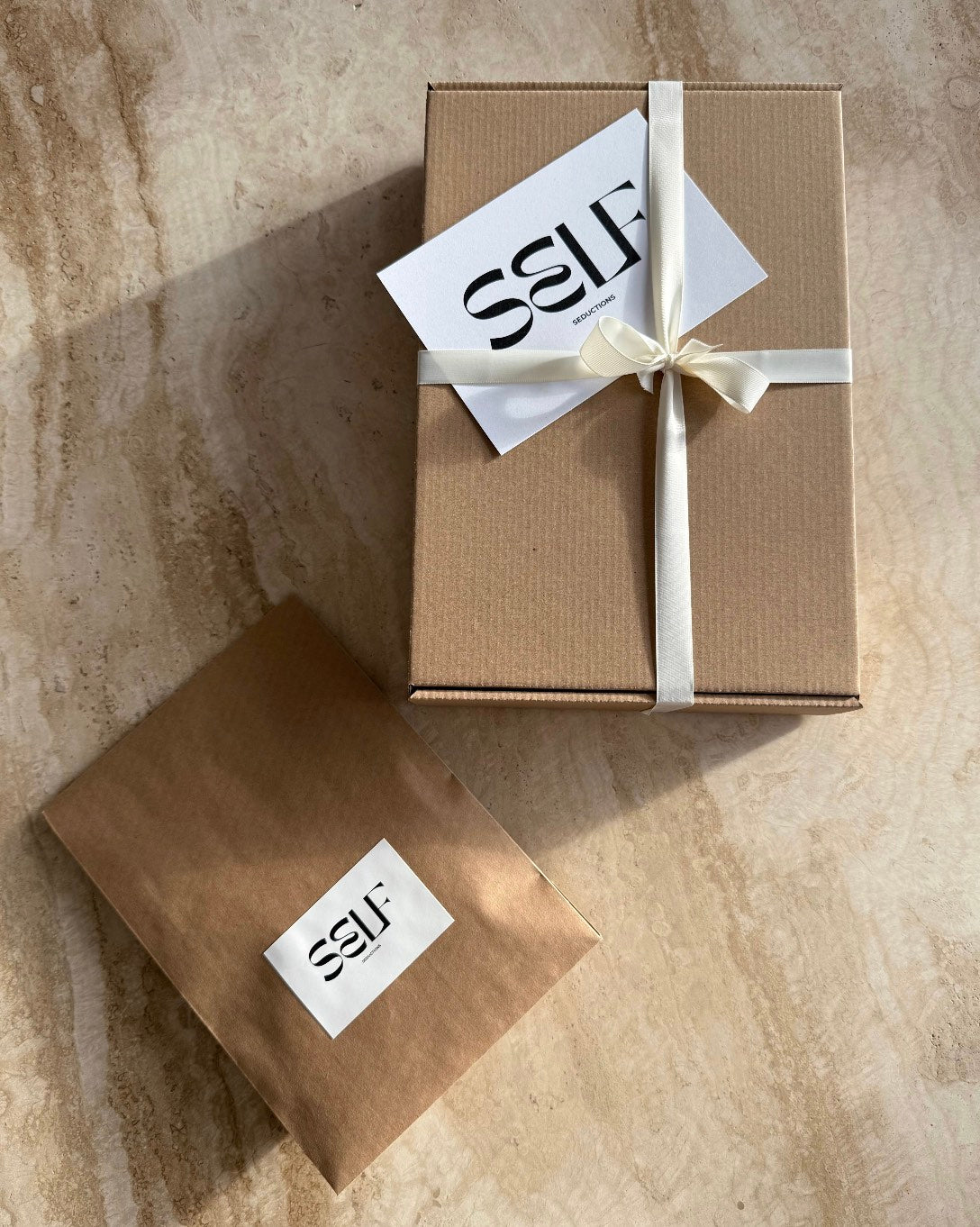 Luxury eco packaging by SELF Seductions – brown mailer with white brand label and tied ribbon on stone surface