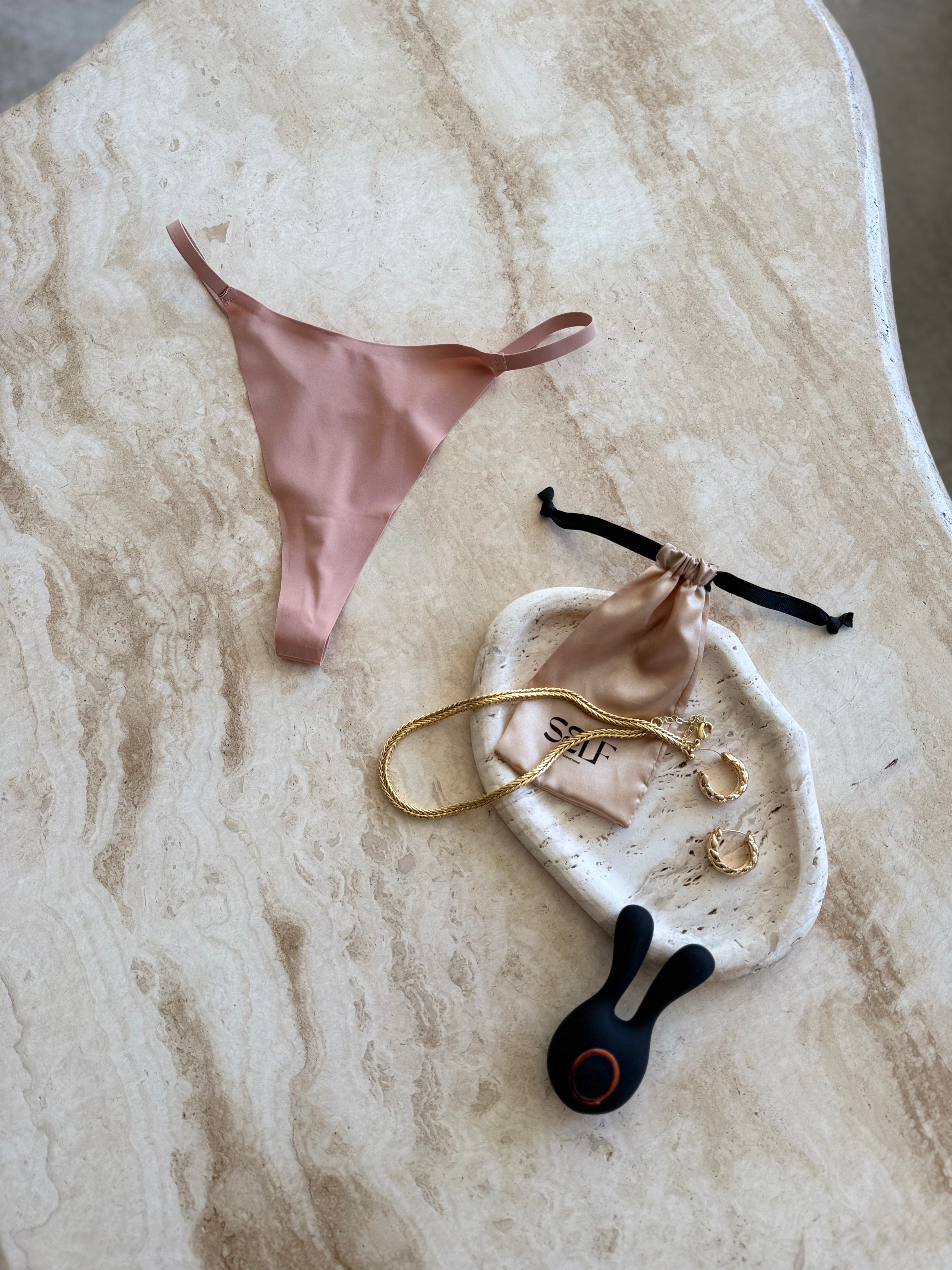 The Kylie Skin Yoga Thong in nude beige – seamless thong with gold jewelry on marble surface – bold yet minimal lingerie still life by SELF Seductions