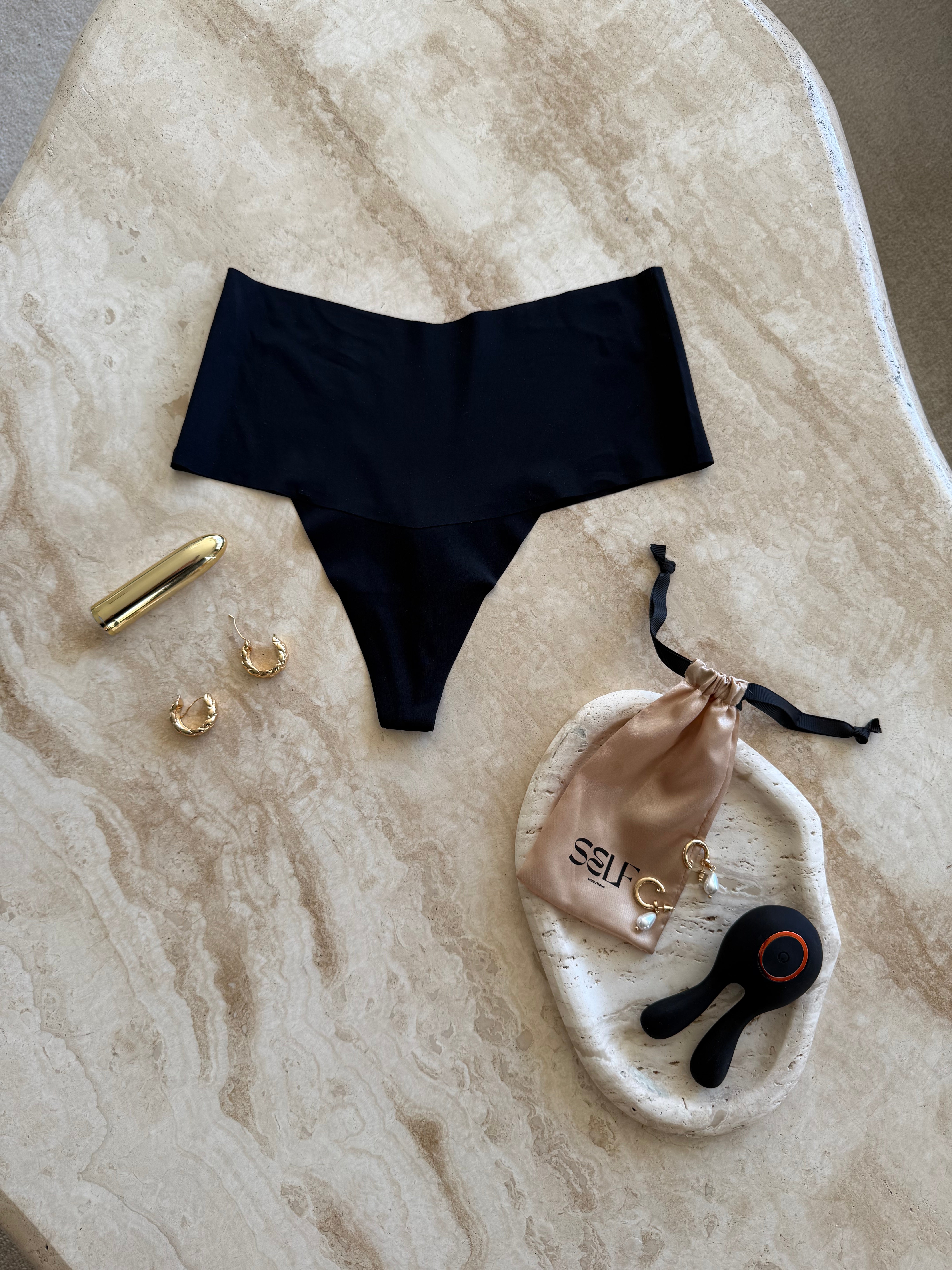 Black seamless high-waist thong with gold jewelry on marble surface – bold yet minimal lingerie still life by SELF Seductions