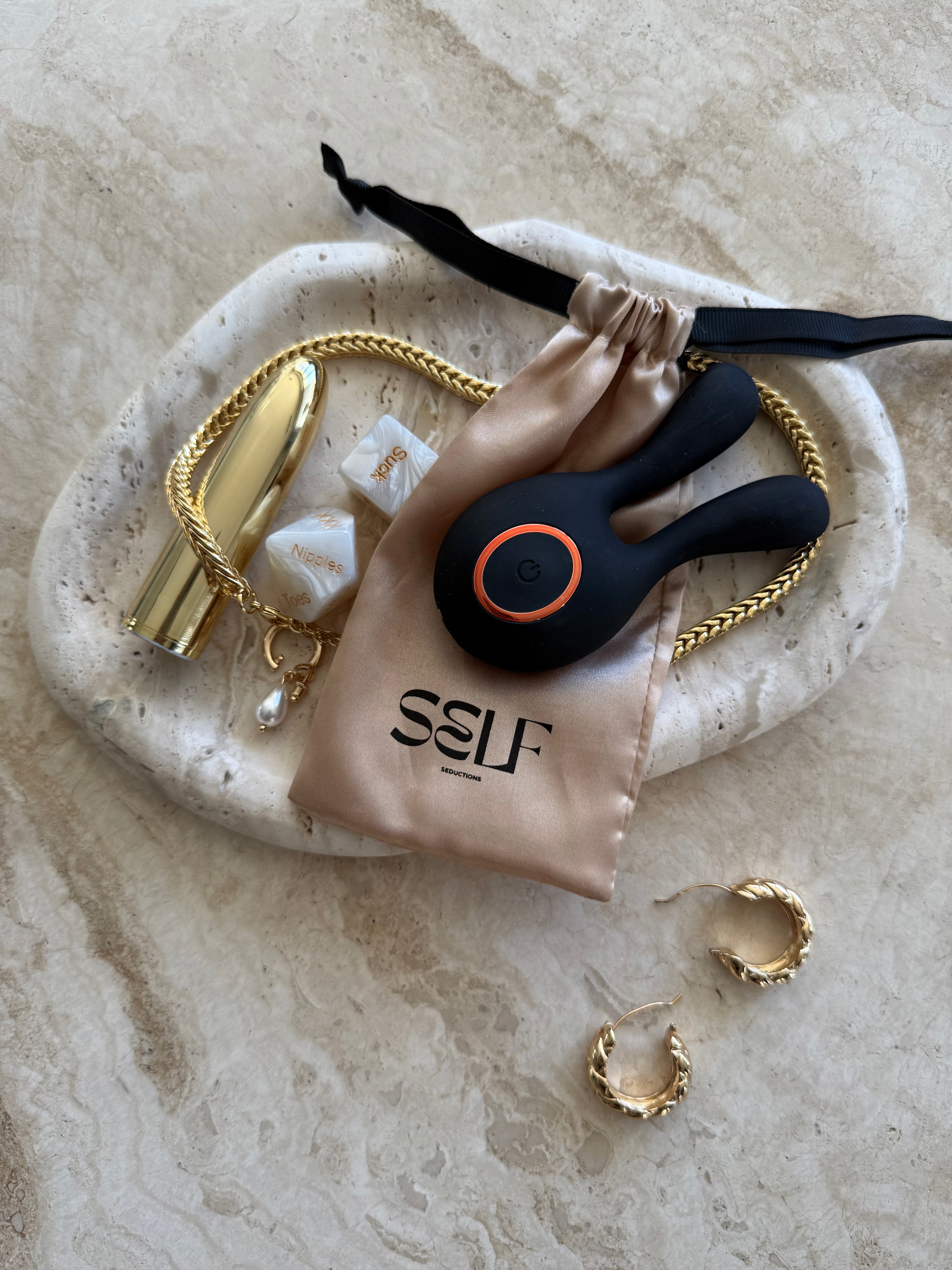 Black twin-ear silicone massager displayed on marble tabletop with gold jewelry and wellness accessory – elegant, sensual still life by SELF Seductions