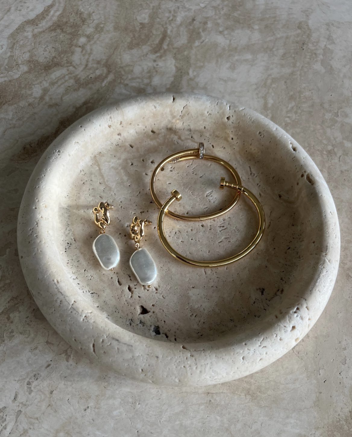 The Nicole Baroque Pearl Drop Earrings in gold with white baroque pearls, styled in a stone bowl with gold bracelets in warm natural light.